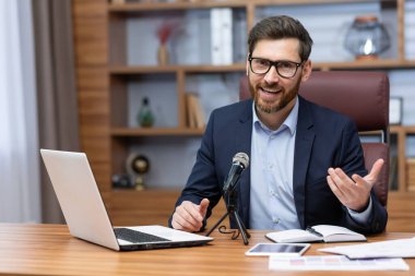 Portrait of successful man inside office, businessman with professional microphone recording online training course and audio podcasts, using laptop sitting at desk working, looking at camera.