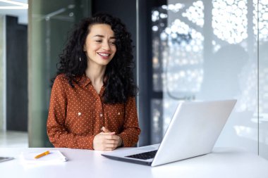 Mutlu ve gülümseyen İspanyol iş kadını dizüstü bilgisayarda yazıyor. Kıvırcık saçlı ofis çalışanı, ofis içi iş yerindeki başarılarından memnun..