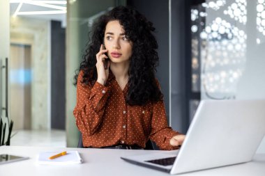 Genç, güzel bir iş kadını telefonda konuşuyor ve pencereye bakıyor. Ofisin içinde iş yerinde dizüstü bilgisayar kullanan mutlu bir İspanyol kadın..
