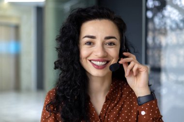 Close up portrait of latin american woman inside modern office with headset for video call, woman smiling and looking at camera, office worker customer support tech helpline.