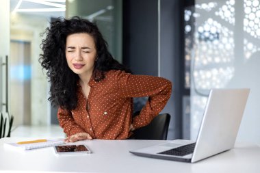 Beautiful Latin American businesswoman working in modern office, worker overtired has severe back pain, using laptop.