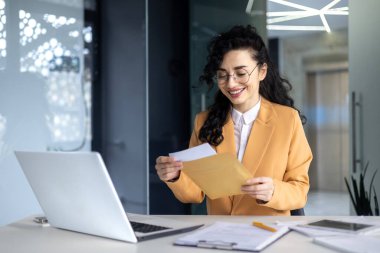 İş kadını bankadan mutlu haber mektubu aldı, kıvırcık saçlı ve gözlüklü İspanyol kadın modern ofis binasında çalışıyor, dizüstü bilgisayar kullanıyor..