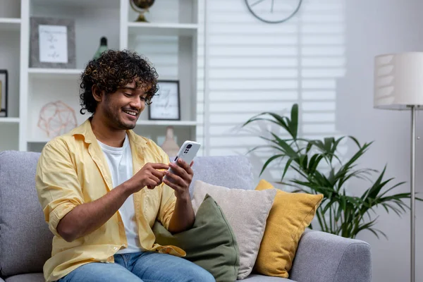 stock image A young Hispanic man is sitting at home on the sofa in the living room, the man is holding a phone in his hands, using an online application on a smartphone, browsing the Internet and typing messages.