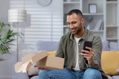 Genç ve mutlu bir Latin Amerikalı adam elinde bir telefonla bir koltukta oturuyor ve aldığı hediyeyle mutlu. Evine teslim edilmiş bir paket..
