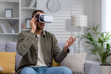 Sanal gözlüklü genç İspanyol adam evdeki kanepede oturuyor ve bir maskeyle çevrimiçi video izliyor. Ellerinle hareket et..