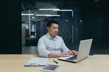 Ciddi ve odaklanmış Asyalı genç işadamı laptopun ve belgelerin arkasında fazla mesai yapıyor..