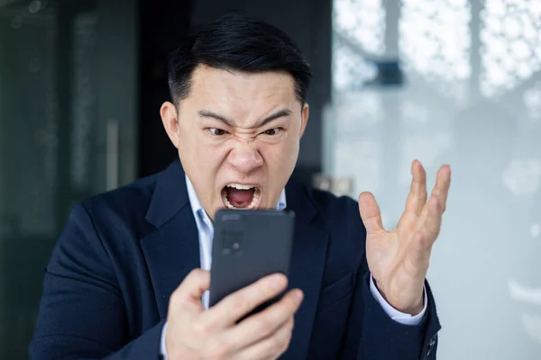 Close-up of angry asian man yelling at phone screen, boss at workplace unhappy with online trading results looking at news on smartphone, businessman investor at workplace.