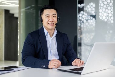 Video görüşmesi için kulaklığı olan başarılı bir ofis çalışanının portresi, Asyalı adam gülümseyen ve ofis içinde çalışan kameraya bakan, müşteri hizmetleri desteği.