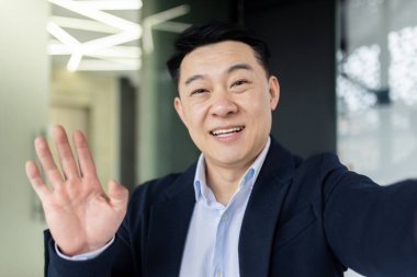 Asian man talking on video call using online communication app on phone, businessman in office taking selfie and smiling at camera, man greeting waving hand while talking with friends.