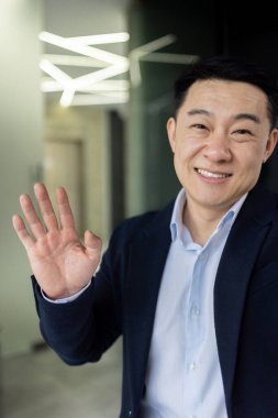 Dikey çekim, Asyalı iş adamı video görüşmesinde konuşuyor, akıllı telefon kamerasına bakıp el sallıyor, selamlama hareketi yapıyor, programcı ofiste çalışıyor. .