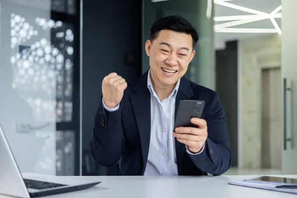 Successful asian businessman received good news notification online, workplace boss holding hand up celebrating successful achievement and winning.