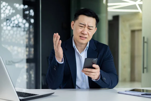 Upset asian man in office at work reading bad news from phone online, businessman near window working inside office, using smartphone