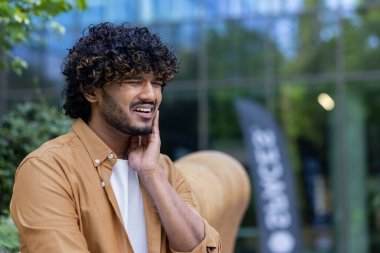 Genç bir İspanyol adam dışarıda bir bankta oturuyor, yüzünü buruşturuyor ve elini yanağına tutuyor. Diş ağrısından muzdarip. Yakın plan fotoğraf..