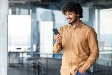 Genç bir İspanyol ofis içinde duruyor, adam telefonu elinde tutuyor, işadamı internet sayfalarına bakıyor ve başvuruları kullanıyor, bir mesaj çeviriyor, bir arama yapıyor, gülümseyen tatmin olmuş bir çalışan..