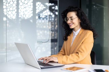 İş yerindeki olgun başarılı iş kadını portresi, kıvırcık saçlı İspanyol kadın dizüstü bilgisayarda çalışıyor, sarı takım elbiseli patron klavyede daktilo kullanıyor, kadın çalışan..
