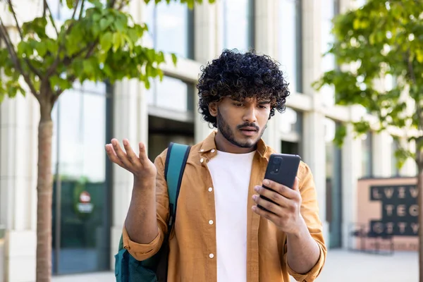 stock image Upset student with backpack reads bad news on phone, man with phone browses internet page on smartphone, opens app, hispanic outside near university campus.