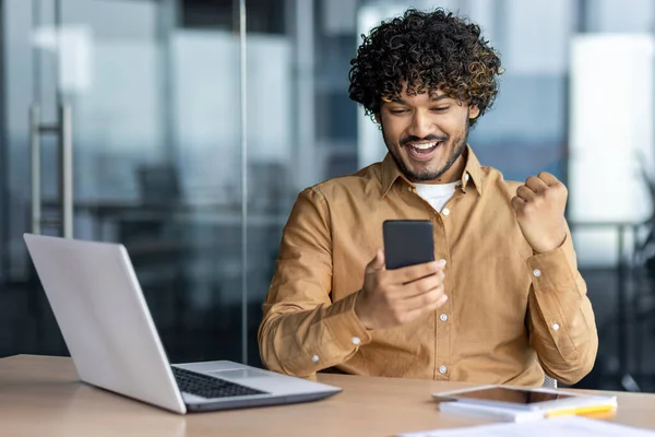 İş yerindeki başarılı Hintli adam zafer ve zaferi kutluyor, iş yerinde telefon işadamını kullanan ofis içinde oturan, akıllı telefon ile çevrimiçi galibiyet haberi aldı.