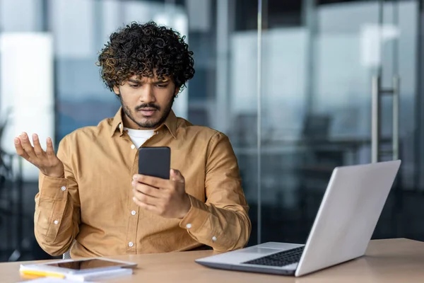 stock image Upset and sad man at workplace inside office, businessman at work received notification message negative, hispanic reading bad news online from smartphone.