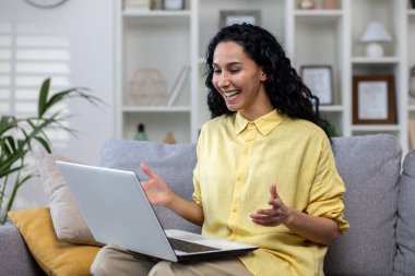 Evde tek başına mutlu bir şekilde konuşan genç bir kadın, video görüşmesi için dizüstü bilgisayarını kullanan İspanyol bir kadın, oturma odasında oturuyor..