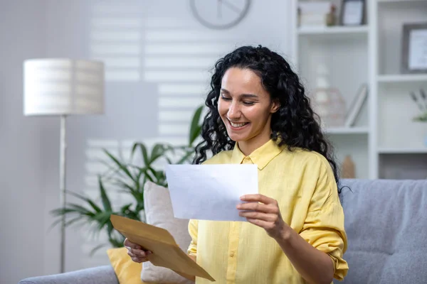 Joyful and happy woman at home received mail envelope with notification, hispanic woman reading good news on paper and happy sitting on sofa in living room .