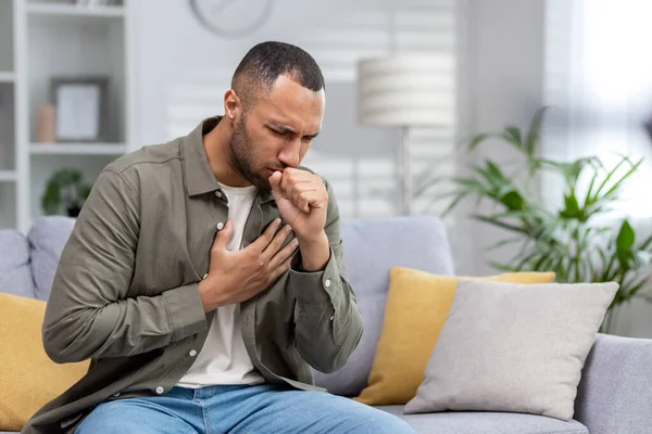 stock image Young African American man sitting on the couch at home and coughing. Suffers from an attack of asthma, allergies. He holds his chest, covers his mouth with his hand.