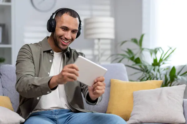 stock image A young Hispanic man plays online games on a tablet at home. Sitting smiling on the couch wearing headphones.