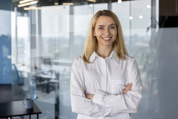 Portrait of a young smiling successful woman designer, engineer, architect standing in a white shirt in the office in front of the camera with her arms crossed on her chest.