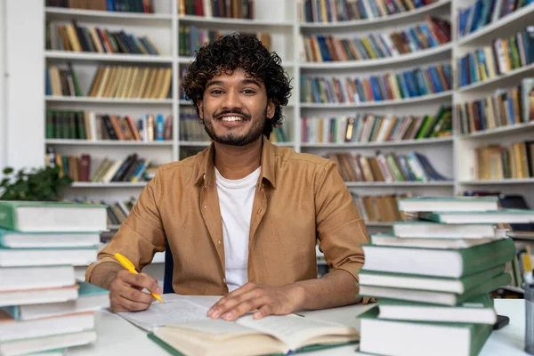 Happy and smiling student studying inside academic library, man smiling and looking at camera doing homework among books.