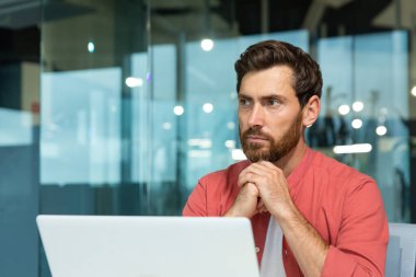 Genç, düşünceli, ciddi bir iş adamının portresi, serbest çalışan, programcı, masada oturan, dizüstü bilgisayarda çalışan, ellerini kavuşturan,.