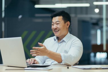 Genç Asyalı erkek işadamı çevrimiçi iş toplantısı, konferans düzenliyor. Ofiste, laptopun önündeki masada oturup video aracılığıyla iletişim kurmak..