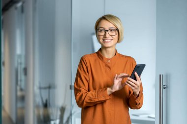 Genç, güzel ve başarılı bir iş kadını koridorda duruyor. Ofiste çalışan, akıllı telefondaki uygulamayı kullanarak gülümsüyor..