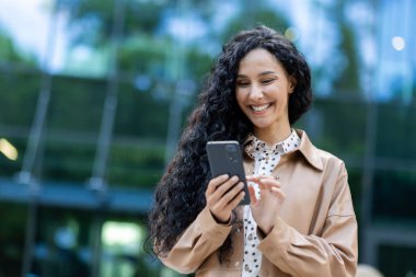 Şehirde yürüyen genç ve güzel İspanyol bir kadın, akıllı telefon uygulaması kullanarak telefonu elinde tutan bir iş kadını, memnun ve mutlu bir şekilde ofis binasının dışında kıvırcık saçlı bir kadın.