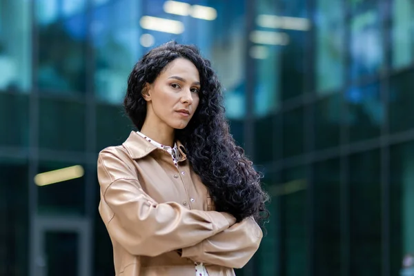Portrait of successful proud woman boss outside office building, hispanic satisfied with work looking at camera with crossed arms, mature businesswoman with curly hair standing and posing.