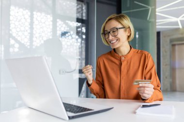 İş yerindeki başarılı ve memnun bir kadın dizüstü bilgisayarla internet üzerinden mutlu banka kartlarını elinde tutan bir iş kadını satın alıyor..