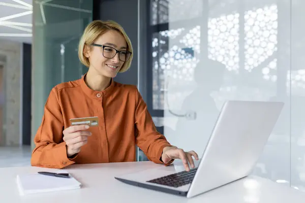 stock image Successful and satisfied woman inside office at workplace with laptop online book so buy business woman holding happy bank credit card in hands.