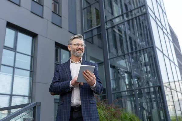 stock image Successful gray-haired businessman smiling and happy, man using tablet computer, senior boss reading news and browsing online pages from outside office building walking around the city.