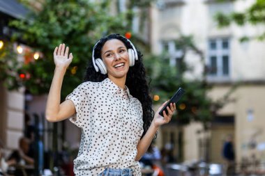 Genç ve güzel Latin Amerikalı bir kadın akşam gezisi için şehre geliyor, kulaklıklı bir kadın müzik dinliyor, dans ediyor ve şarkı söylüyor, telefonunda bir radyo uygulaması kullanıyor.