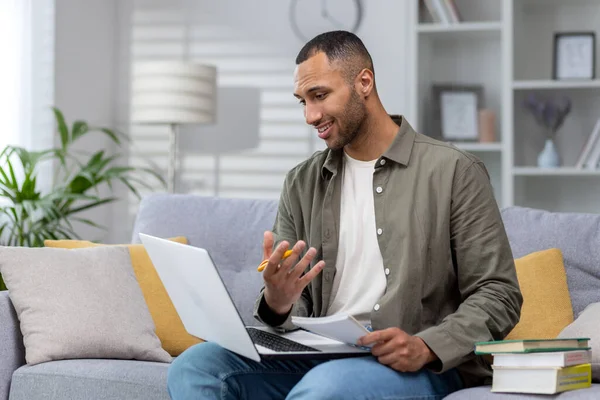 stock image Young African American male teacher sitting on sofa at home with laptop on lap, books and notebook. Teaches dissonance online. Reads a lecture, explains on a video call to the camera.