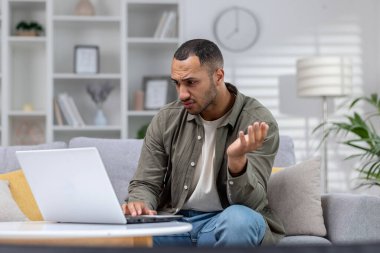 Endişeli Afrikalı Amerikalı adam dizüstü bilgisayar ekranına bakıyor. Evdeki kanepede oturuyor ve ellerini açıyor. Cihazla ilgili sorunlar, kötü haberleri okumak, bir mektup almak.
