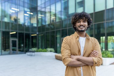 Kampüsün dışında kollarını göğsüne kavuşturup kameraya gülümseyen genç, kendine güvenen, yakışıklı ve zeki Hintli erkek öğrencinin portresi..