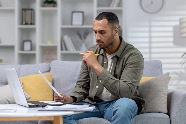 stock image Worried and serious young African American man working at home with documents and bills. He sits huddled on the sofa in front of the laptop and holds papers in his hands.