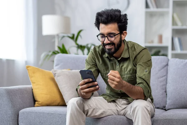 Happy and satisfied with achievement results hispanic at home reading online win notification in online application on phone, man smiling holding hand up triumph gesture sitting sofa in living room.