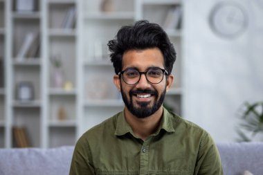 Oturma odasındaki kanepede oturmuş gülümseyen, gülümseyen ve kameraya bakan İspanyol bir adamın yakın plan portresi..