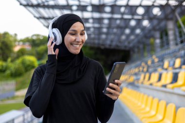 Stadyumda elinde kulaklıklar ve telefonlarla tesettürdeki Müslüman sporcu kadın, aktif egzersiz ve spor yaparken kadın, dışarıda müzik dinleyen ses kitapları ve podcast uygulamaları..