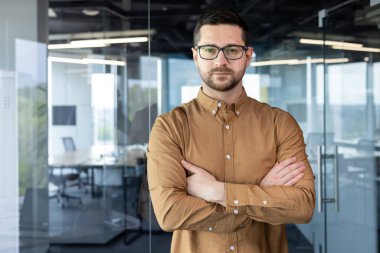 Kendine güveni tam genç bir işadamının portresi, yönetici direktör, bir ofis binasında duran kurucu. Kollarını göğsünde kavuşturur ve kameraya ciddi ciddi bakar..
