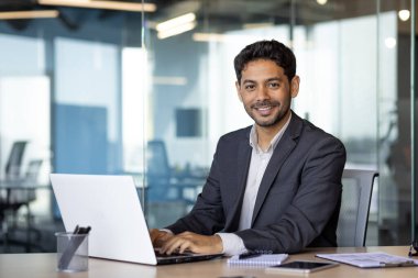 Genç Arap iş adamının portresi, ofiste otururken gülümseyen ve kameraya bakan adam, iş yerindeki takım elbiseli patron dizüstü bilgisayar kullanıyor..