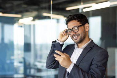 Başarılı yatırımcı ofis içinde çalışır, iş adamı takım elbiseli iş adamı pencere kenarındaki telefonu kullanır, adam gülümser ve akıllı telefondan iyi haberler okur.