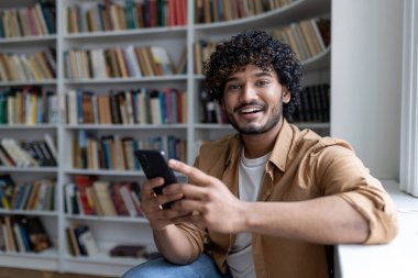 İspanyol asıllı genç bir öğrencinin portresi, elinde telefonla gülümseyen ve kameraya bakan bir adam, üniversite kampüsünde halk kütüphanesinde eğitim görüyor..