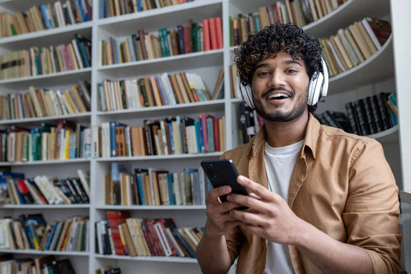 stock image Young hispanic student inside public library studying, smiling and looking at camera, man holding phone, using study app, with headphones among shelves.