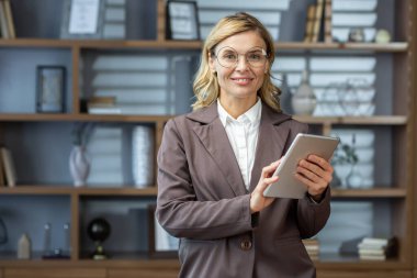 İş yerinde çalışan olgun, deneyimli bir kadın, elinde tablet bilgisayarla iş kadını portresi, patron gülümseyip kameraya bakıyor..